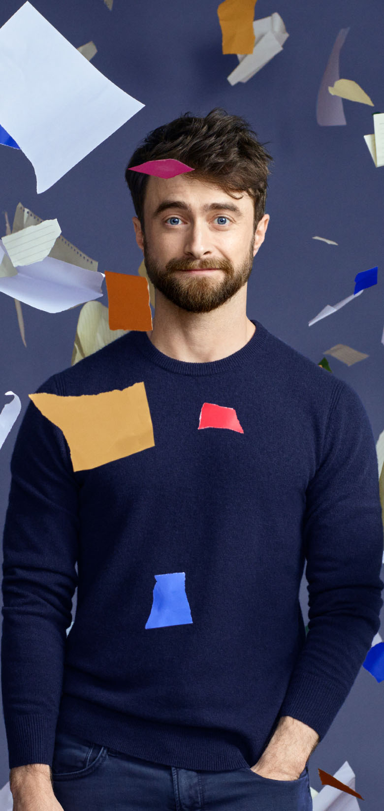 Daniel Radcliffe stands amidst floating, colourful paper fragments against a dark background, smiling.